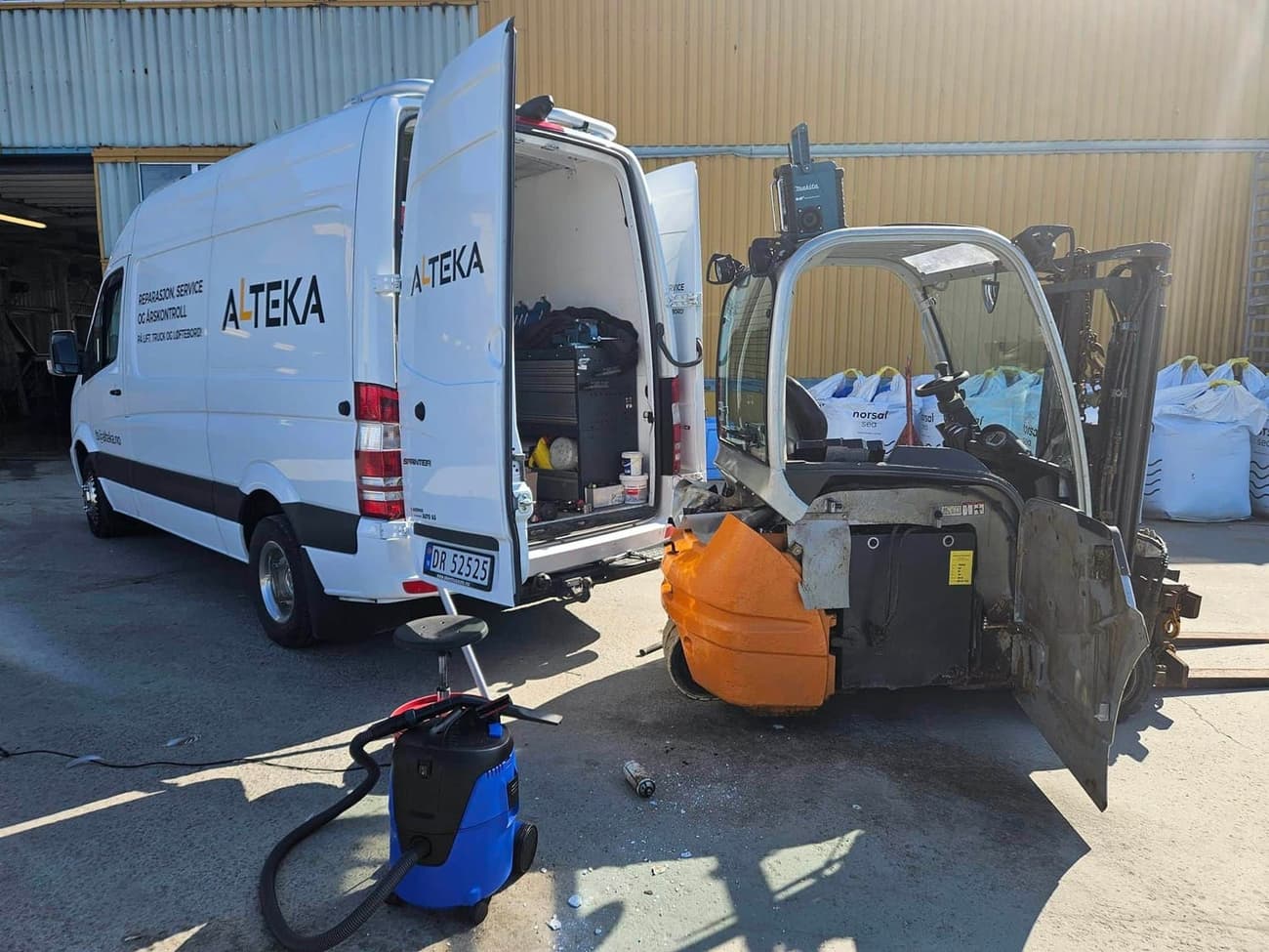 En truck delvis demontert foran servicebilen utenfor verkstedporten, med verktøy og støvsuger på asfalten